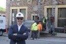 A man in a business suit stands with a hard hat near construction work going on behind him.