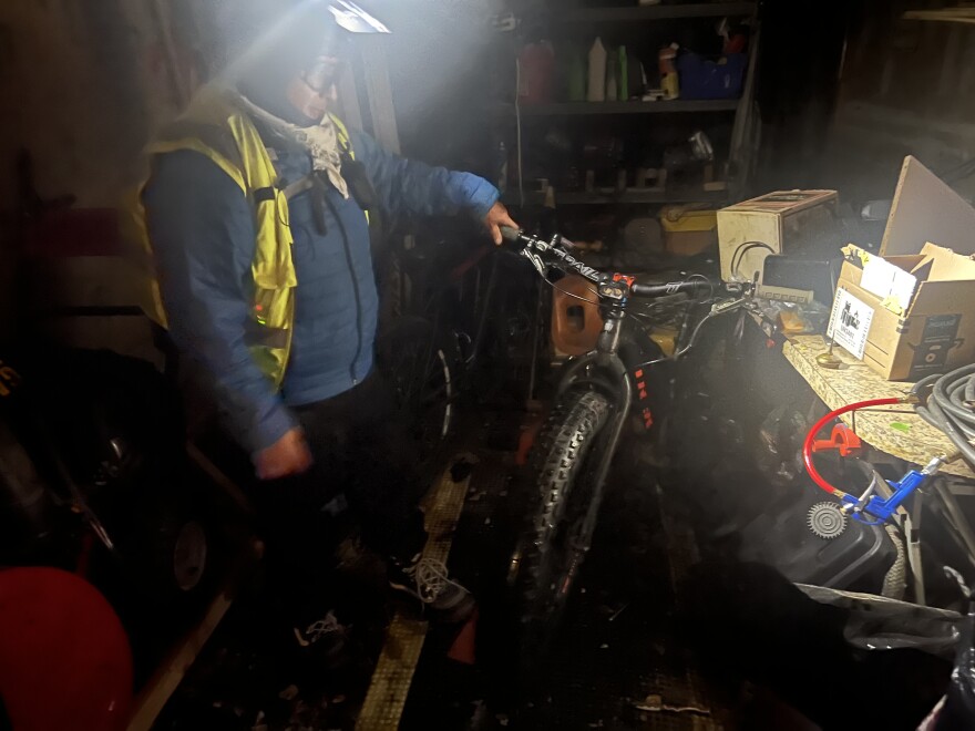 A man dressed in cold weather clothing holds the handlebar of his fat tire bike inside a garage illuminated by a headlamp attached to his bike helmet.