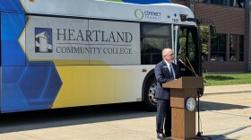 A man stands speaking at a podium, with a large bus behind him. The Connect Transit bus has a hawk on it and says Heartland Community College on the windows.