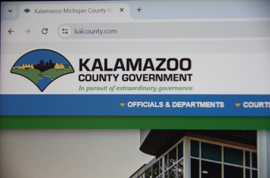 A computer monitor displays the Kalamazoo County Government Website. The URL at the top of the page reads "kalcounty.com." The page is predominantly white, though the addition of blue and green text and banners livens up the page.  A graphic next to the page title shows the Kalamazoo River running between to hills populated by yellow trees. Black buildings standing against a clear blue sky can be seen in the background of the logo. Below the page title sits Kalamazoo County's motto in green italicized text. It reads, "In pursuit of extraordinary governance."