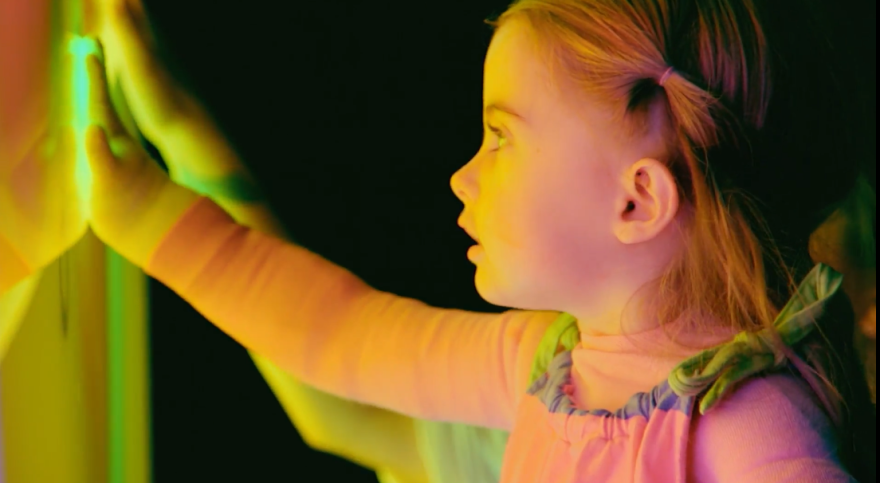 Rowan, a small child with blond hair and a pastel-colored outfit, visits a light installation that lights their face with a golden glow in a still from the documentary "Matter of Time."