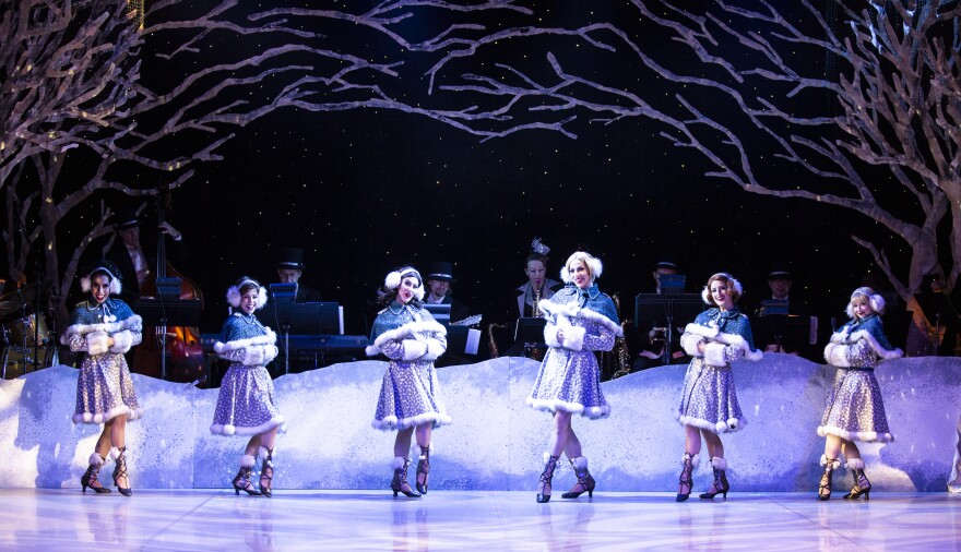 Six dancers dressed in blue-ish dresses with a fur trim and heels stand center stage in front of a background of snow and the jazz band.