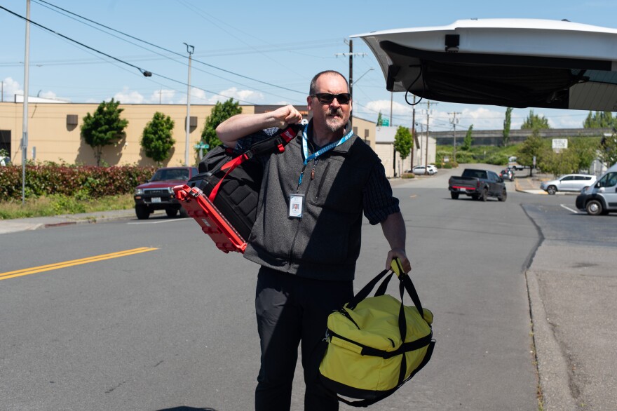 A man grabs two large duffle bags, one black and one yellow, from the trunk of a vehicle on a sunny day. Cars are parked on the street in the background. 