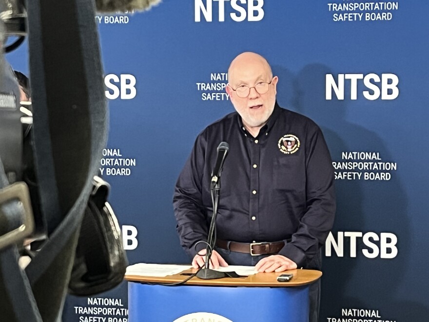 This is a photo of NTSB Board Member Tom Chapman speaking at a lectern in front of a single microphone during a press conference in Cheektowaga. Chapman is older man with a beard, no hair and glasses. He's wearing a navy blue button-up emblazoned with the NTSB's logo on it. A television camera can be seen in the left side of the image. 