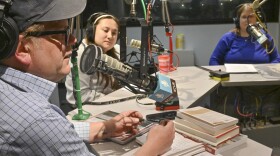 Three people are seated inside a radio studio. They are sitting behind microphones. The man at left, is talking and holding a pen in both hands with a small stack of books in front of him.