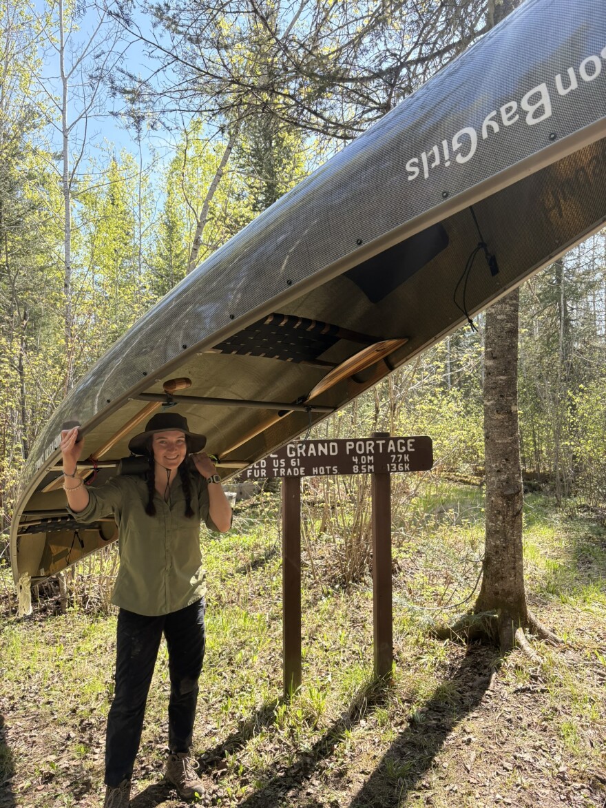 A woman wearing a broad rimmed hat with a chin strap is holding a canoe above her head. Behind her a sign reads "Grand Portage," which was the beginning of the group's journey.