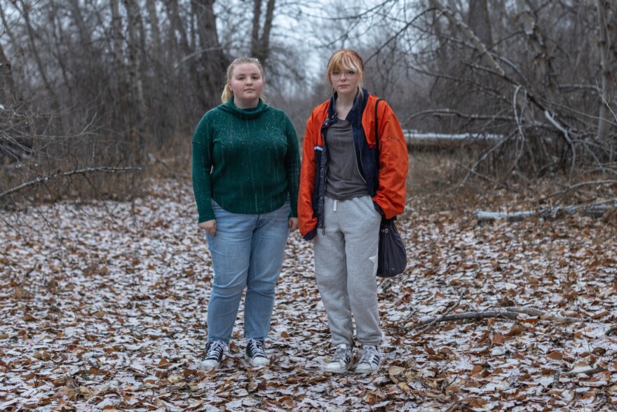 Two teens - one with blonde hair wearing blue jeans and green sweater. The other teen is wearing gray sweats, glasses, a gray t-shirt, an orange jacket with a black bag on their shoulder. Both are standing in forest clearing covered in brown leaves.