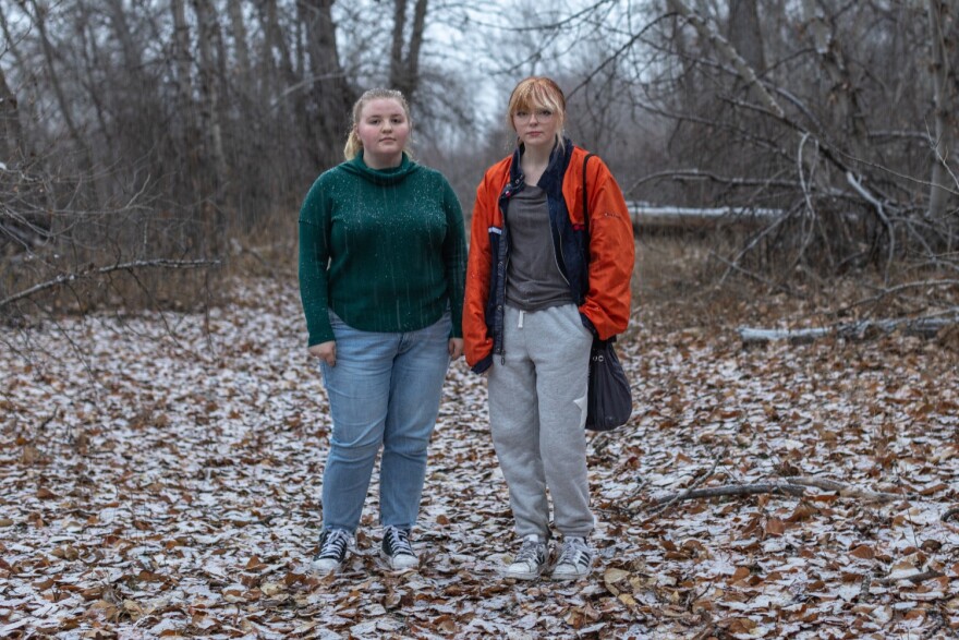 Two teens - one with blonde hair wearing blue jeans and green sweater. The other teen is wearing gray sweats, glasses, a gray t-shirt, an orange jacket with a black bag on their shoulder. Both are standing in forest clearing covered in brown leaves. 