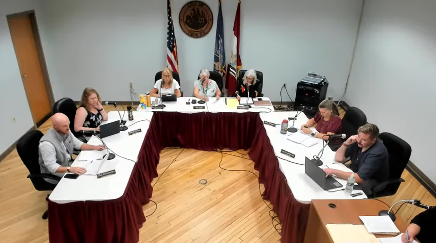 The seven members of the Troy City Council are sitting in a U-shaped arrangement of tables. There's some flags in the background. 