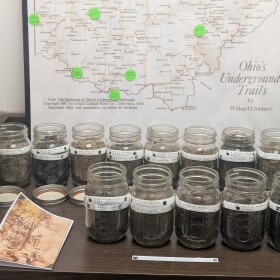 14 jars filled with dirt sit on a shelf in front of a map of the Underground Railroad stops in Ohio. Each jar is labeled with a man's name and a city name. 