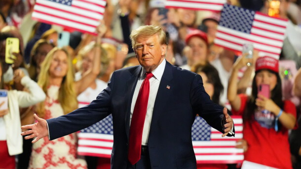 President Donald Trump stands with his arms outstretched. A crowd behind him waves American flags.