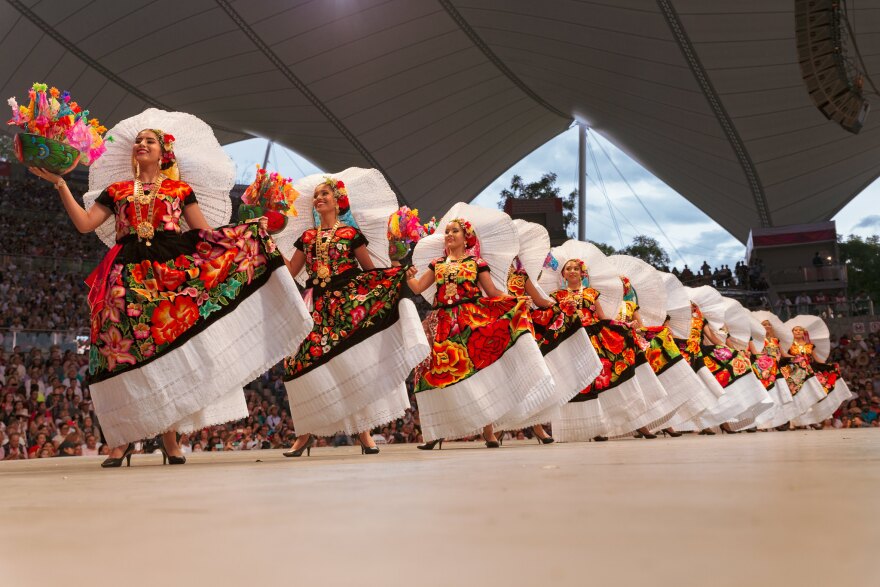 A row of women carrying flowers and wearing colorful floor-length dresses and large white headdresses. 