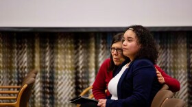 A woman puts her arm around a young woman. Both are seated in chairs facing the front of a room. 