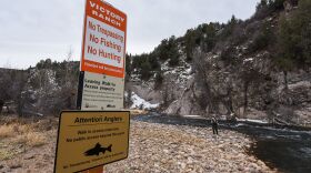 Signs saying, "no trespassing, no fishing, no hunting," "leaving walk-in access," and "attention anglers," are hung on a post in the foreground, with a section of the Provo River and a fisherman in the background.