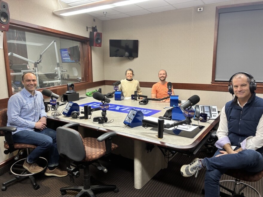 Four people wearing headphones sit at a table in a radio talk studio: a man at left foreground has short dark hair and is wearing a white and blue plaid button-down shirt, jeans and brown shoes; a man at right foreground has short dark hair and is wearing a navy puffy vest, white button-down shirt, jeans and sneakers; a woman at left background has grey hair in a ponytail and is wearing a cream sweater and black turtleneck; a bald man at right foreground has a brown beard and is wearing an orange t-shirt.
