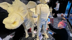 people in warm clothing carve a boy and cow out of butter inside a large refrigerated display cooler.