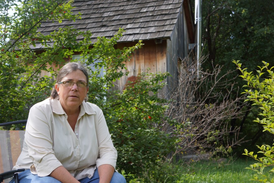 A woman in glasses sitting in a chair looking into the camera with  greenery and a cabin in the background