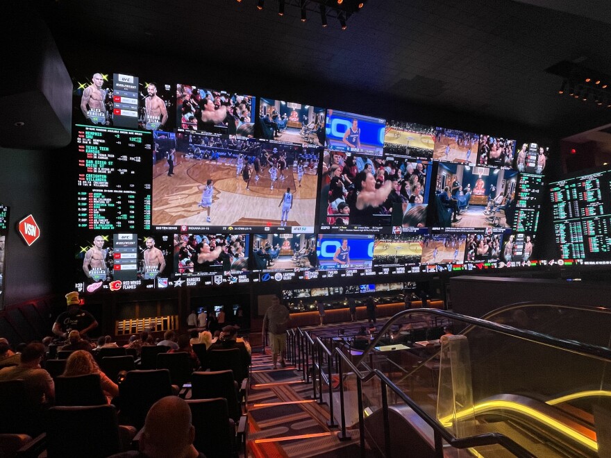 A large panel with multiple TV screens shows various views of a basketball game and data related to it while people sit in a movie theater-like setup watching the screen.