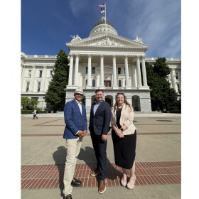 Three people stand in front of the California State Capitol on a sunny day. 