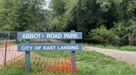 This image shows a dark green sign with white text that reads, "Abbot Road Park City of East Lansing." There is orange fencing around the sign because of ongoing construction. The sign sits on a patch of green grass. Behind and next to the grass is are dirt trails leading into the park. There is green foliage in the background.