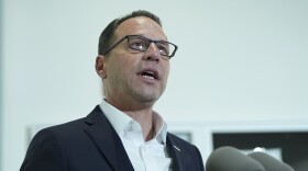 A man in a suit with glasses speaks into multiple microphones at a podium.