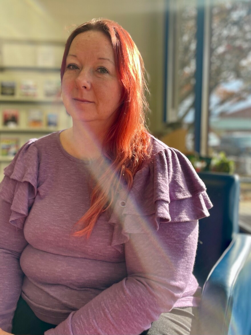 Sarah Smith works as a medical assistant at an urgent care in Christiansburg. She has red hair, draped over her left shoulder. She's sitting near a window with bright orange light coming through the window shining on her hair. The look on her face could be relaxed, tired, content, or patient. There is a sense from seeing her that she has an inner strength that keeps her going. 