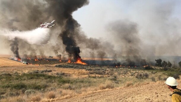 A small white and red plane flies low over a wildfire consuming brush and dry grass in a field. BLM trucks and firefighters are also around the perimeter of the fire. On firefighter is in the lower righthand corner of the image with a yellow shirt and white hardhat. 