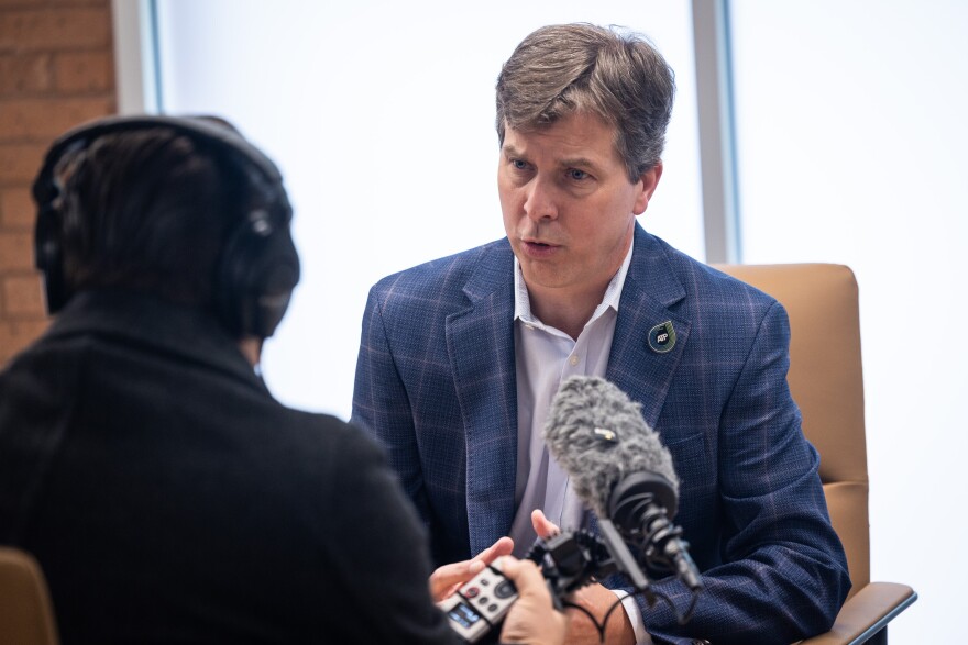 Austin Transit Partnership Director Greg Canally doing an interview. He's wearing a blue suit and speaking into a microphone while seated at a desk.