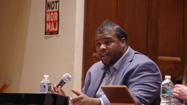 A man in a light blue suit sits at a desk and speaks into a microphone. 