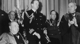 A black and white photograph showing Winston Churchill wearing academic robes seated on the right with dignitaries behind him and President Harry S. Truman standing on the left, also in academic robes looking at Churchill and applauding.