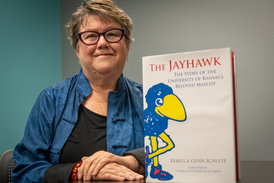 A woman sits at a table with her hands folded in front of her. Beside her, on the table, stands a large, coffee-table book called "The Jayhawk, the Story of the University of Kansas's Beloved Mascot."