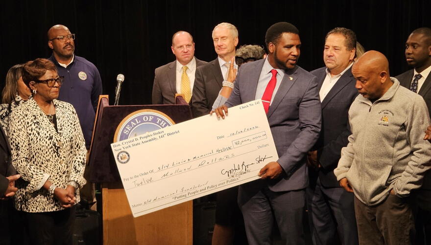 5/14 Memorial Foundation Executive Director Chad Houston, holding check, reacts to the foundation receiving $12 million in state funding. The money was presented by Assembly Majority Leader Crystal Peoples-Stokes, far-left, and Assembly Speaker Carl Heastie, far-right in gray jacket.