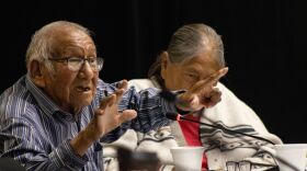 An Oglala elder sits at a table wearing a blue striped button down shirt. He is holding his hands up as he speaks while a woman sits beside him and listens. 