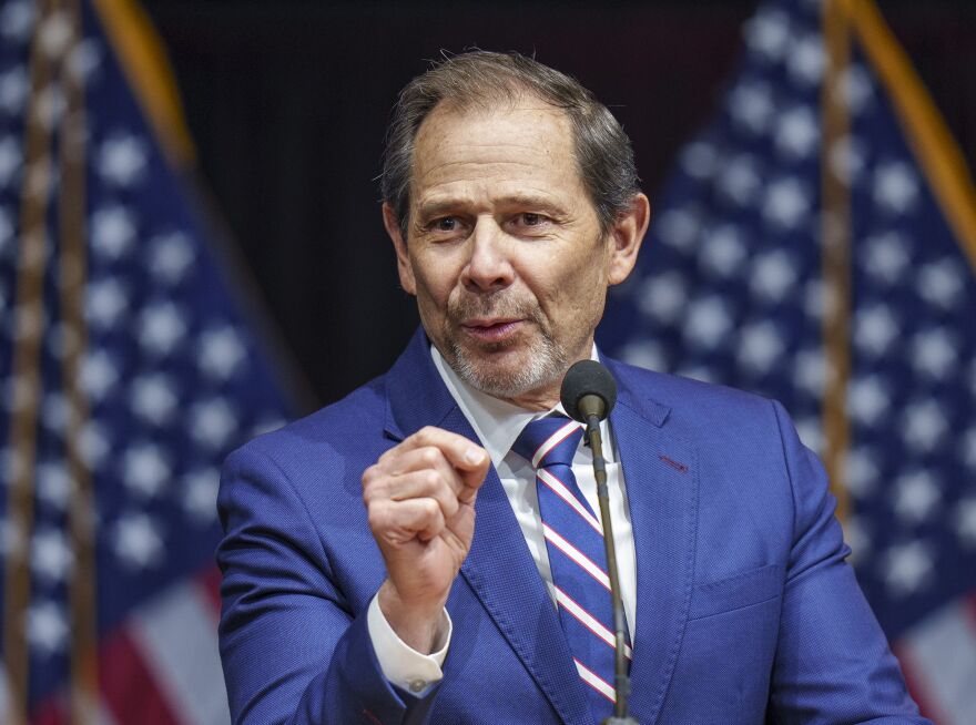  U.S. Rep. John Curtis wears a blue suit while he speaks behind a microphone, with American flags in the background.