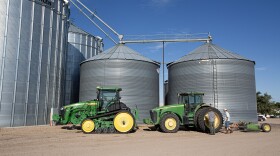 Two men stand next to a huge wheel of a green tractor. This tractor and another green tranctor sit in front of two medium tall metal grain silos and two very tall metal grain silos. The ground is brown dirt and the sky is blue.