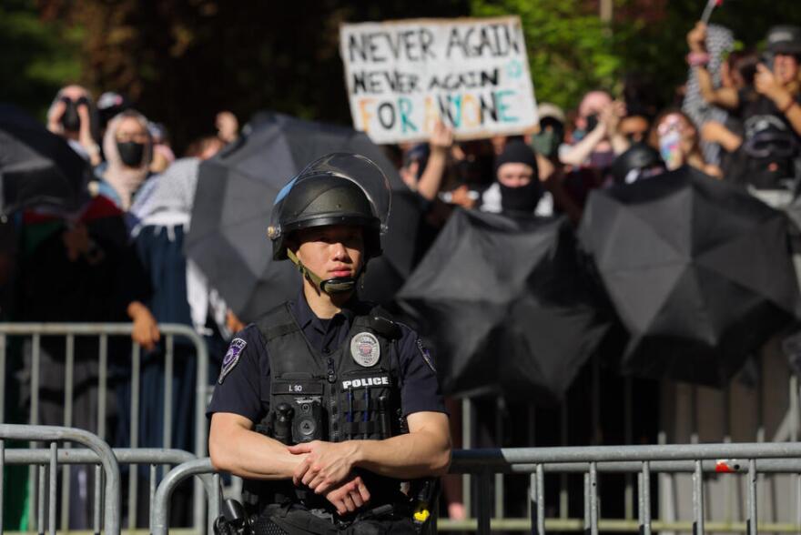 A police officer in a uniform, vest and helmet stands in front of barricades. A couple of feet away in front of a barricade, protesters stand with masks, umbrellas, and signs.