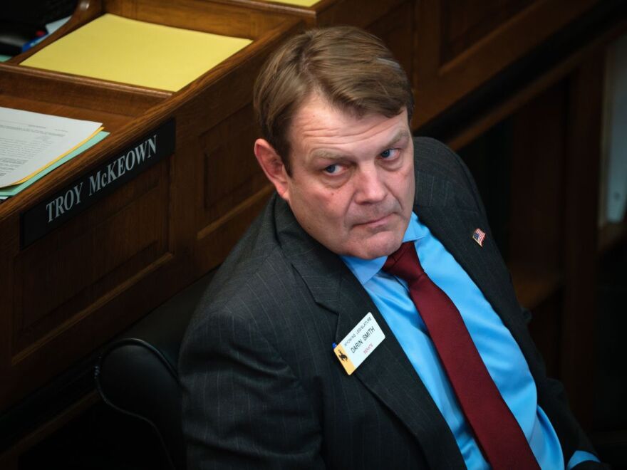 A man wearing a light blue dress shirt, maroon tie and dark blazer glances to the left of the camera while sitting in the Wyoming capitol building.
