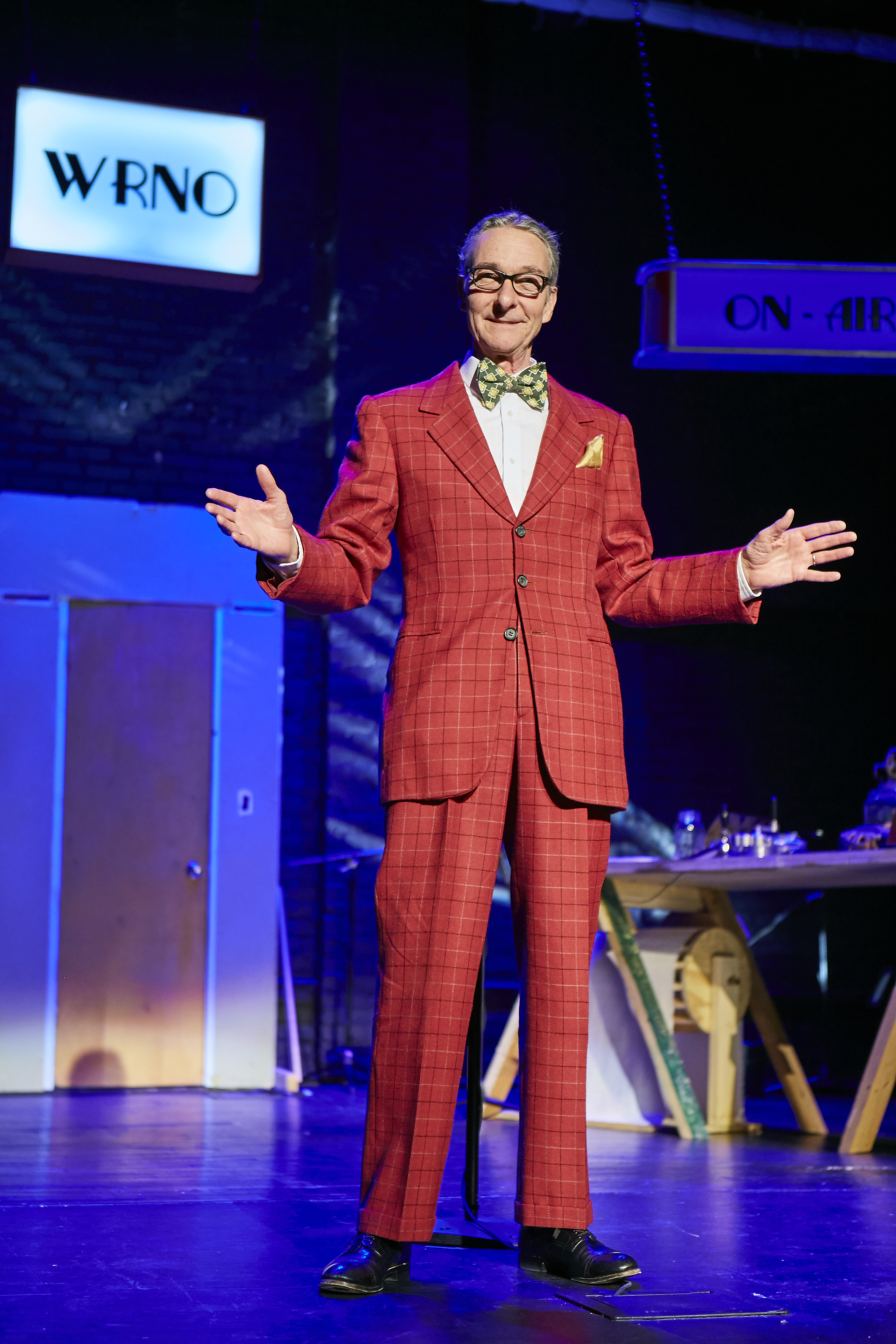 A performer in a bright red period suit stands at a microphone with his arms open