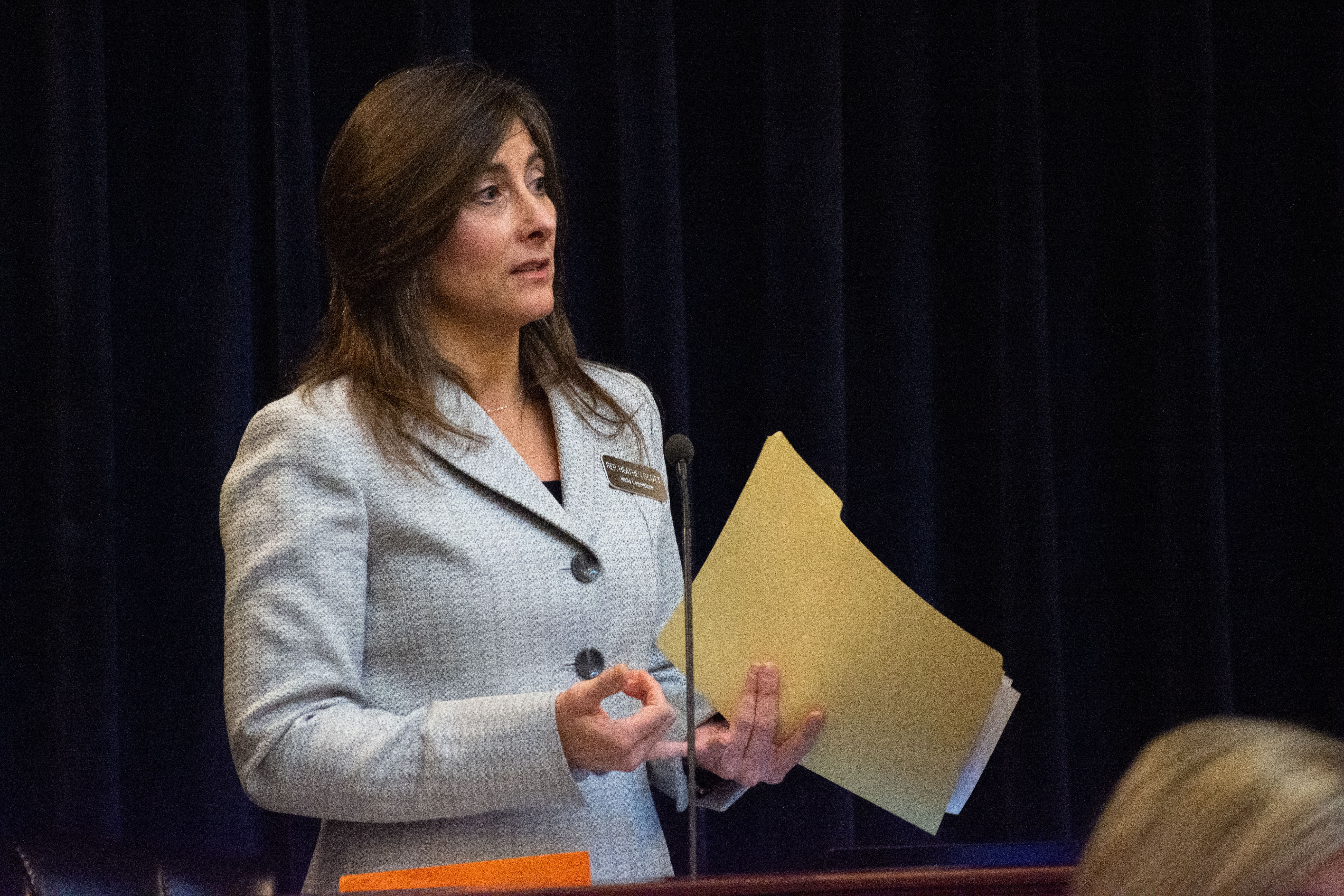 A woman stands behind a small microphone with a file folder in her hand.
